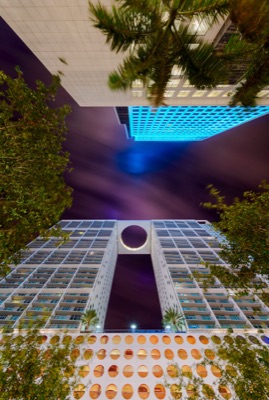 Night-time upward view between illuminated high-rises and tree branches towards a purple sky, with a large opening near the top of one building.