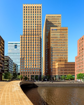 View across water toward the Amsterdam Symphony high-rise towers by De Architekten Cie, featuring textured brick facades under a clear blue sky.