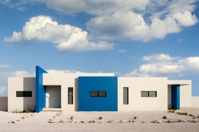 Front of Coco Loco by McNiff & Obersi, with flat roofs, white and cobalt-blue walls and louvred windows facing a gravel plot under a partly cloudy blue sky.