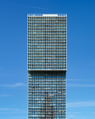 Graphic architectural photograph: A straight-on view of a tall modern skyscraper with a gridded glass facade and an overhanging upper section against a clear blue sky — Rotterdam.