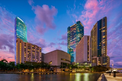 High-rise buildings and skyscrapers in Downtown Miami illuminated along the waterfront at sunset, under a blue sky with pink clouds.