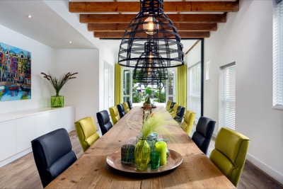 A long wooden dining table lined with black and chartreuse chairs beneath black cage pendant lights, set beneath exposed ceiling beams with a view toward the patio.