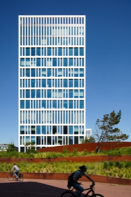 A modern white high-rise building with a slatted facade stands against a clear blue sky, overlooking terraced planters with weathered steel edges and cyclists in the foreground.