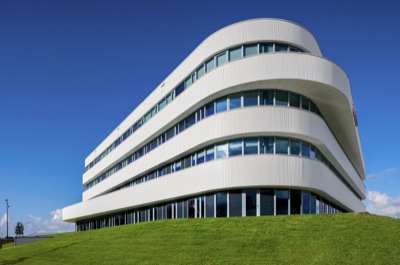 Low-angle view of the Extra Verde building by MVSA, featuring curved white horizontal facade panels and continuous window bands atop a green grassy hill against a clear blue sky.