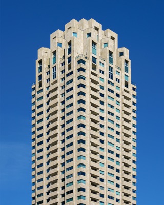 Graphic architectural photograph: Upper portion of a tall stone-clad skyscraper with a stepped geometric crown, numerous windows, and balconies set against a clear blue sky — Rotterdam.