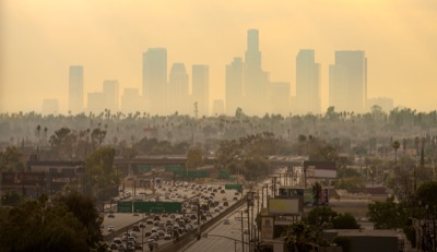 Distant downtown Los Angeles skyscrapers rise above haze, with a busy multi-lane freeway carrying traffic amidst palm trees and buildings in the foreground.