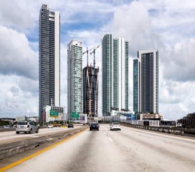 View from a multi-lane highway with moving cars heading toward a modern skyline of tall skyscrapers, including one under construction with cranes, under a cloudy sky.