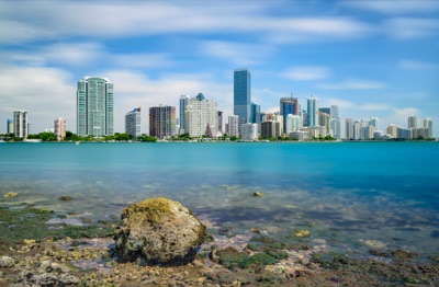 View of the Miami skyline across calm turquoise water, featuring a rocky shoreline with a prominent stone in the foreground under a blue sky.