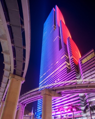 A modern skyscraper illuminated at night in blue, purple, and red lighting, seen from below past elevated transit tracks and palm trees.