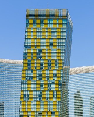 Graphic architectural photograph: A leaning modern skyscraper with a patterned yellow and reflective glass facade, flanked by curved high-rise glass buildings under a clear blue sky — Las Vegas.