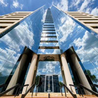 Low-angle view looking straight up at a modern skyscraper with reflective glass walls mirroring white clouds and a bright blue sky.