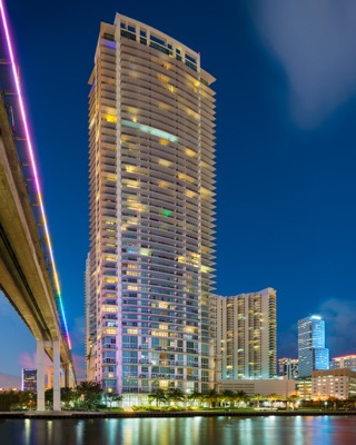 A tall illuminated skyscraper stands by the water at dusk, next to an elevated bridge with colorful lights and reflections across the river.