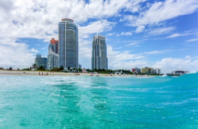View from the turquoise ocean waves towards the sandy shoreline of South Beach, featuring high-rise buildings including Continuum and Portofino under a blue sky with clouds.
