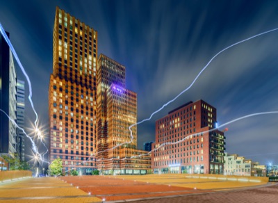 Long-exposure night photograph of modern illuminated brick high-rise buildings and a paved square, crossed by wavy glowing light streaks.