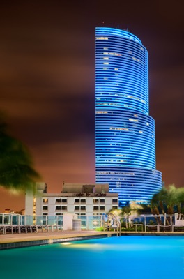 Night view of the Miami Tower glowing in bright blue light behind a swimming pool with deck chairs and palm trees.